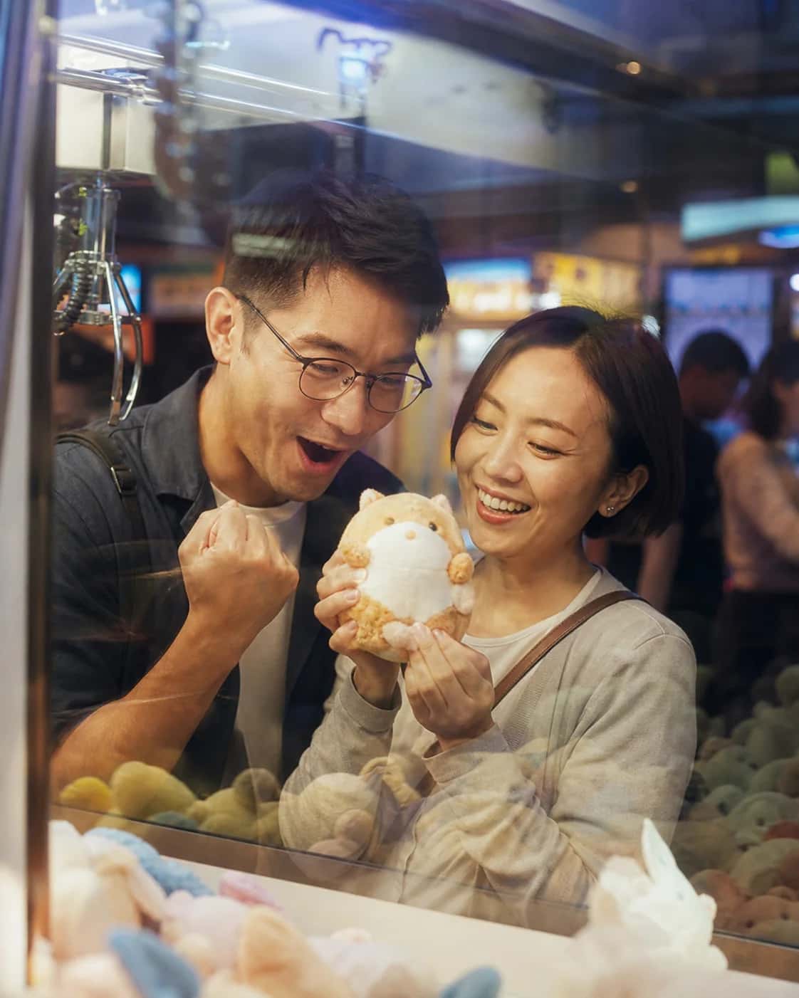 Illustrative brand scene of two adults holding a plush toy beside an arcade claw machine; not a member photo or testimonial.