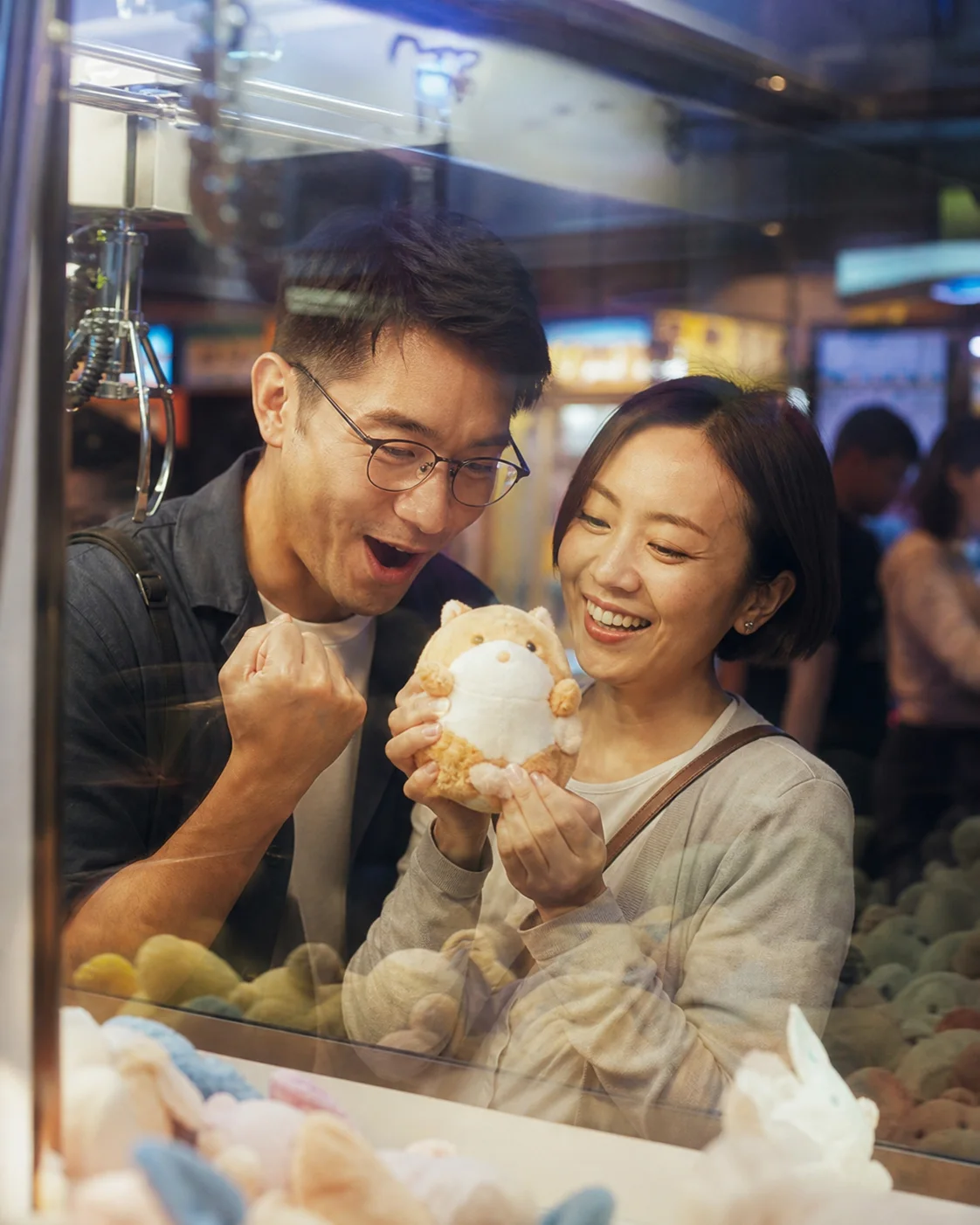 Illustrative brand scene of two adults holding a plush toy beside an arcade claw machine; not a member photo or testimonial.